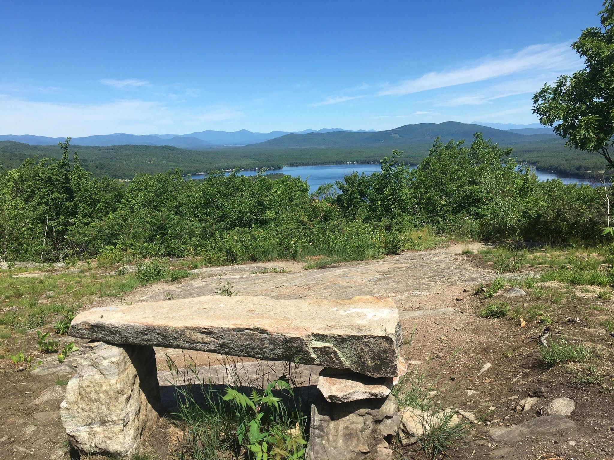 View of Province Lake from the summit of Province Mountain. In the foreground, a rough-hewn stone bench and bright green foliage of deciduous trees. In the distance, low mountains. Above, blue sky with a few white clouds.