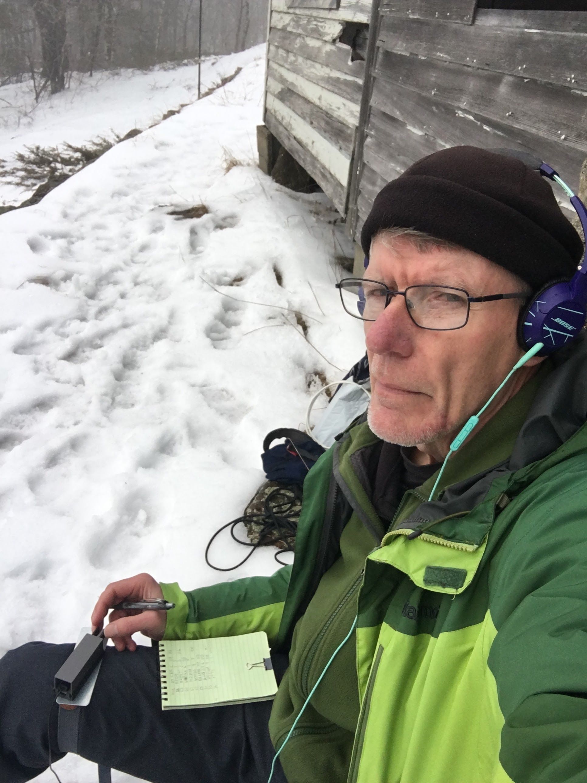 A man in winter wear sits outdoors leaning on a clapboard-sided shack. Or N1AIA pauses sending CW for a selfie.