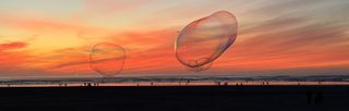 Soap bubbles float over a wide beach colored orange at sunset. Far away, people walk the water's edge.
