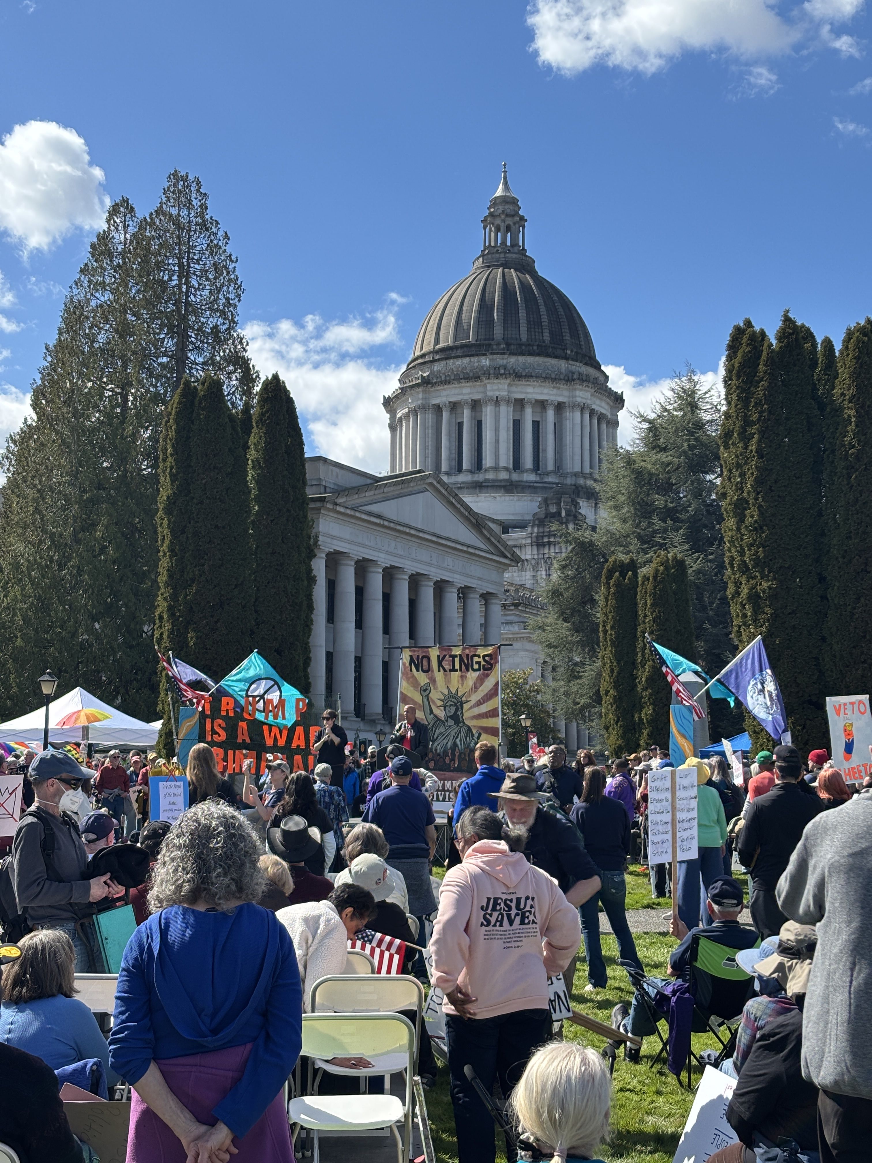 In-the-audience perspective of the No Kings event stage with the Capitol dome in the background. Dozens of people are facing away from the camera while a speaker addresses the crowd, A.S.L. translator by his side. Protest signs are in abundance.